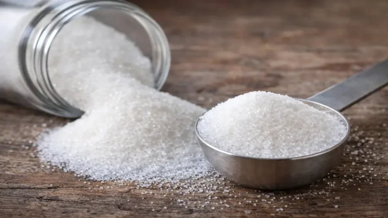 Sucre semoule en grains fins dans une cuillère avec bocal renversé sur surface en bois, mise en scène réaliste en cuisine